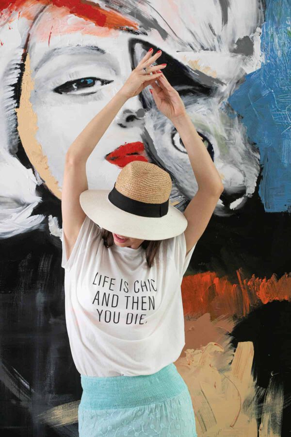 תמונת אווירה - קריאייטיב ליין - דוגמנית עומדת על רקע ציור גדול עם חולצה לבנה עליה כתוב (באנגלית) : " החיים הם שיק ואז אתה מת"