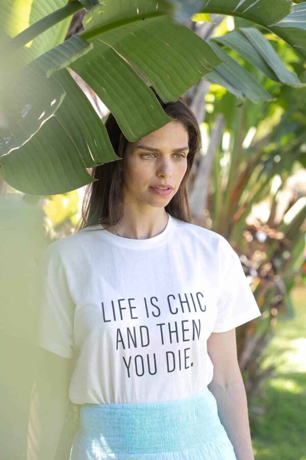 תמונת אווירה - קריאייטיב ליין - דוגמנית עומדת על רקע עץ ירוק עם חולצה לבנה עליה כתוב (באנגלית) : " החיים הם שיק ואז אתה מת"